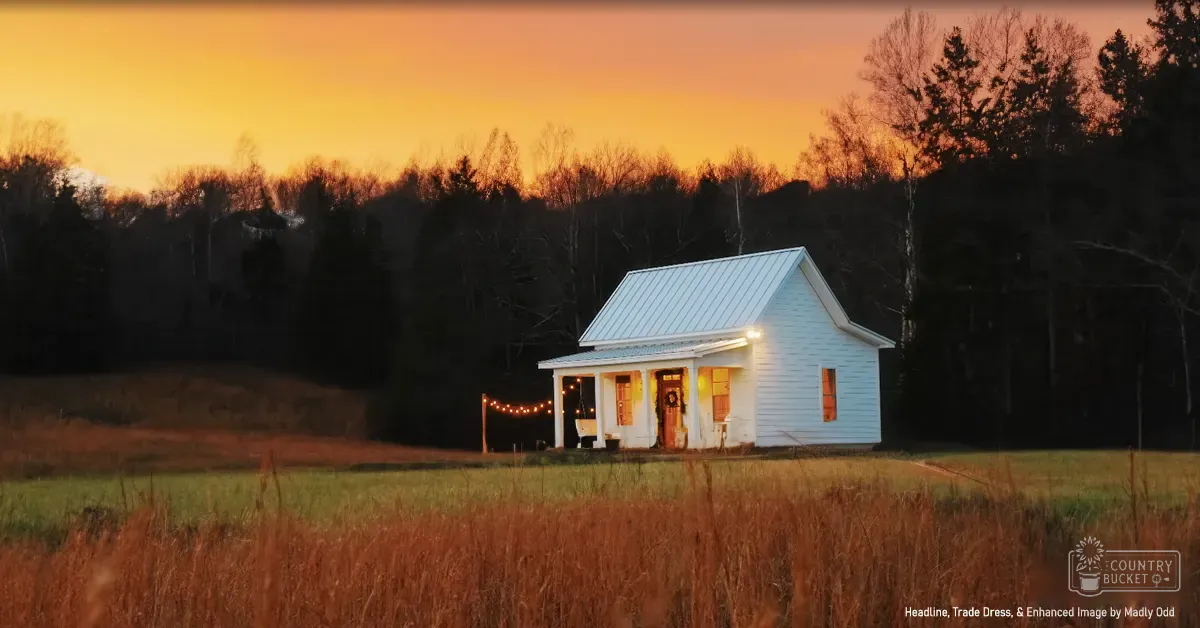 A small white house with a gabled roof, porch adorned with string lights, surrounded by grassy terrain in late autumn or early winter at sunrise or sunset.