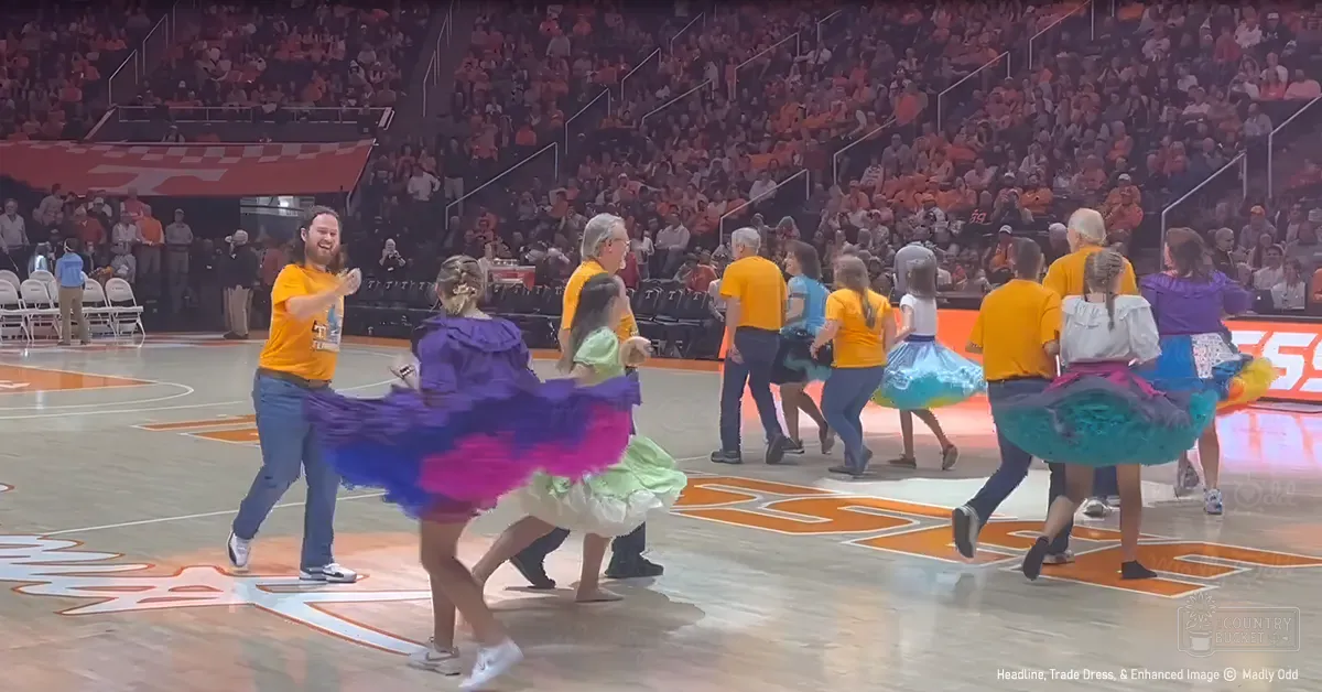 A group dance performance at an indoor sports arena with bright orange t-shirts and tutu skirts on young girls among spectators wearing team colors.
