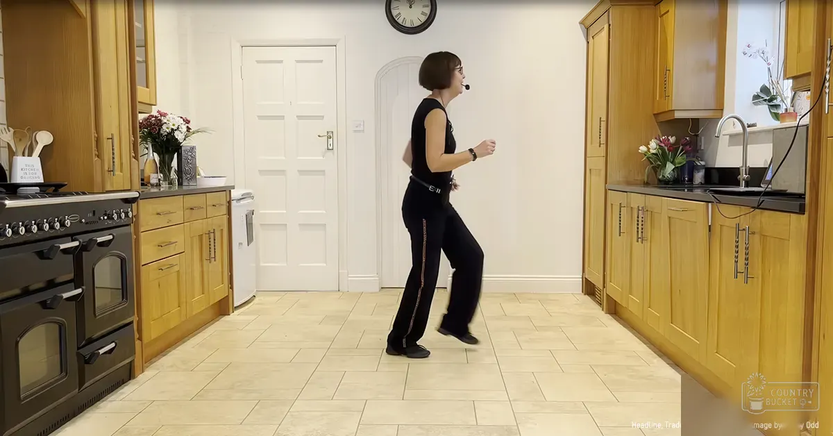 A young adult female dances in a kitchen, wearing black pants with thin gold stripes, a sleeveless black top, and black shoes. The scene includes wooden cabinets, tiled floor, flowers on countertops, clock above the door, and various kitchen items.