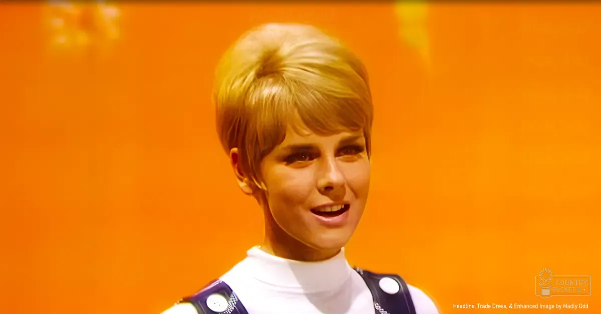Young adult female in a white turtleneck sweater with dark strap, set against bright orange background. Studio or performance space with mid-20th century style garment and 'Headline, Trade Dress, & Enhanced Image by Madly Odd' text visible on clothing.