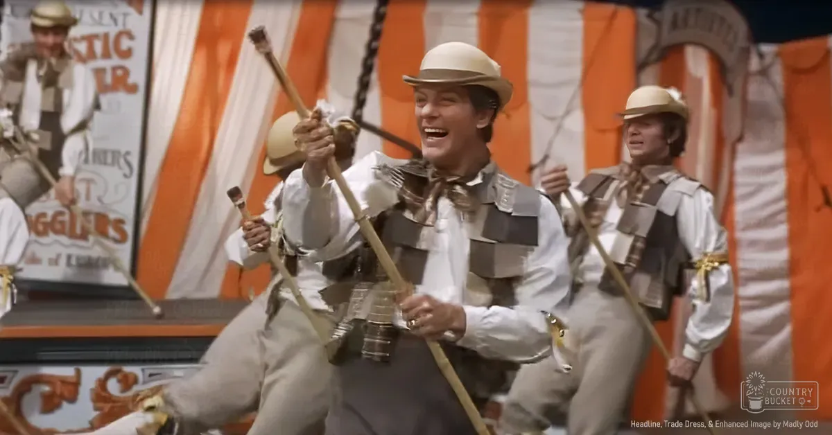 A lively dance scene inside a circus tent with performers wearing white shirts and beige vests, holding drumsticks.