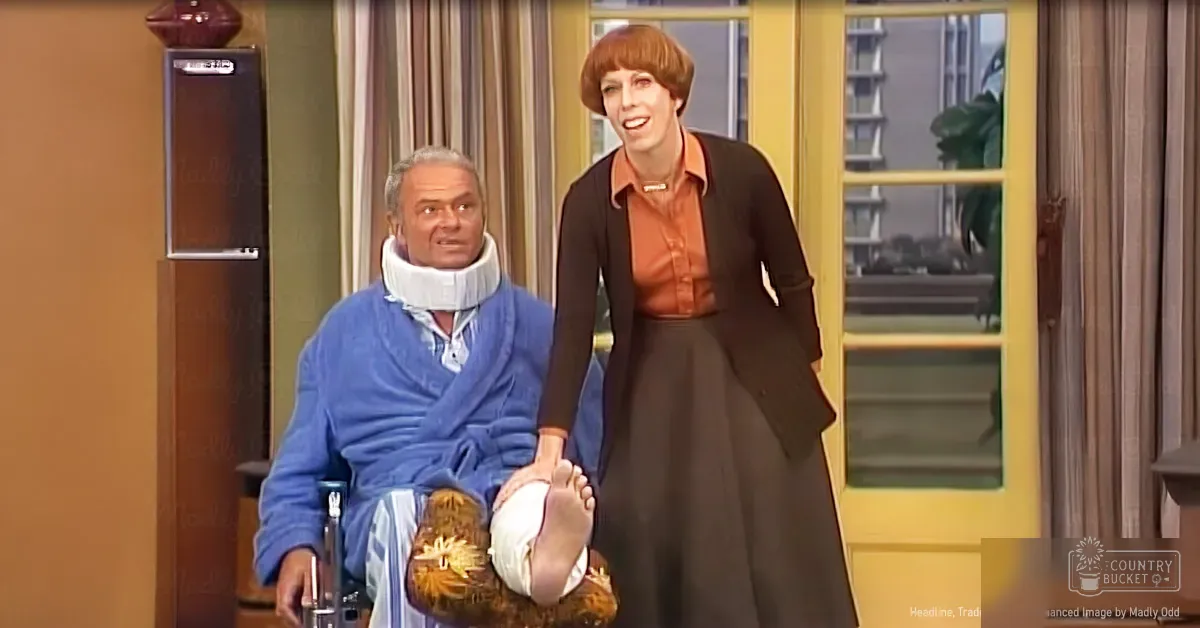 A middle-aged man in a blue patterned robe with a white cast and crutches, seated in a wheelchair indoors near an adult female wearing a black high-neck dress and orange blouse under natural daylight.