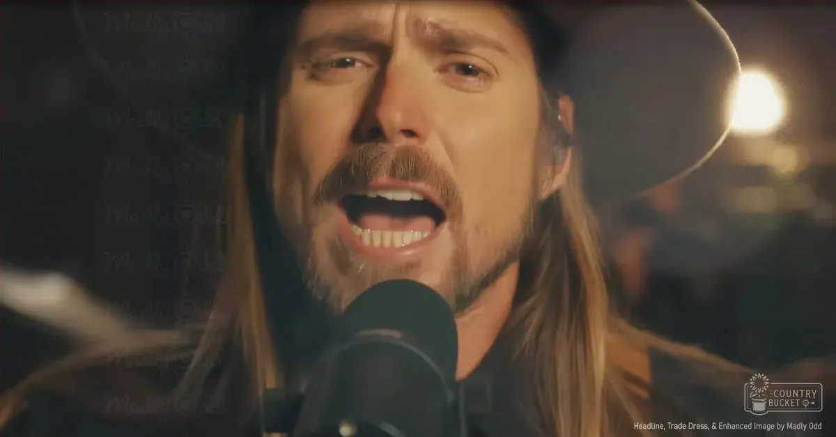 An adult male singer wearing a dark shirt with a light vest underneath and a black hat, performing indoors with a microphone and text overlay reading 'Madly Odd'.