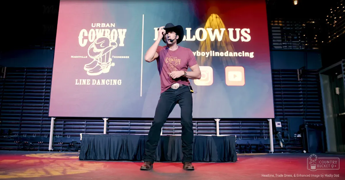 An adult male singer in a maroon t-shirt with brown boots, performing at an event in Nashville, Tennessee. The stage has red carpet and displays 'Urban Cowboy Line Dancing' along with social media handles.