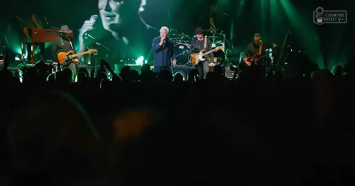 A band performs on stage with green lighting, audience in shadow, and a large screen showing a singer.