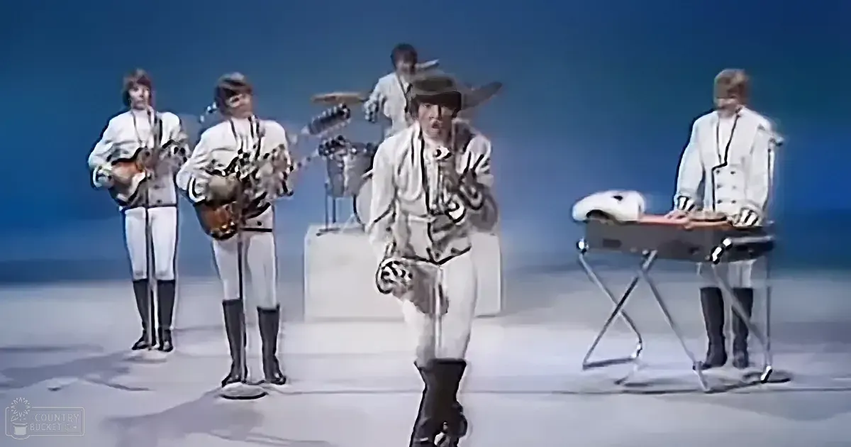 A band in white outfits performs on stage with a blue background, playing instruments.