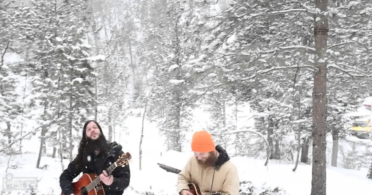 Two adult males in a snowy forest setting, one holding an acoustic guitar and wearing a dark jacket over a lighter shirt or sweater; the other in casual attire with an orange beanie. The scene is illuminated by natural sunlight filtering through snow-covered trees.