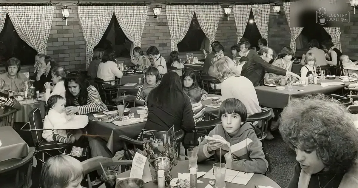 A boy sits at a table in a busy restaurant, surrounded by other diners, with a checkered curtain backdrop.