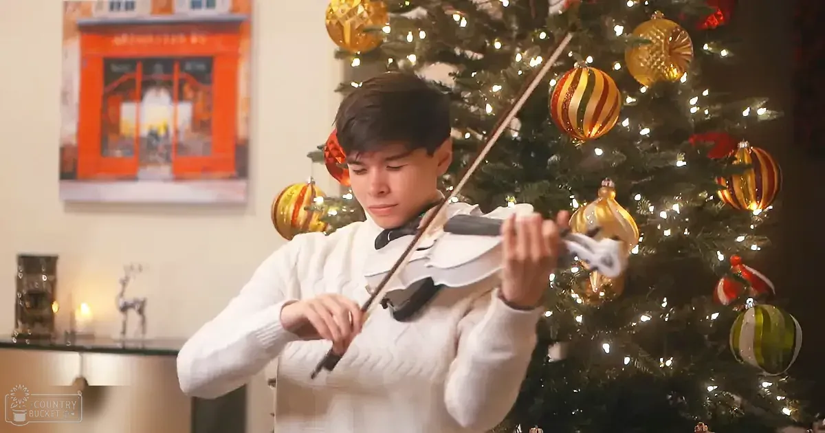 A boy in a white sweater plays the violin in front of a decorated Christmas tree.