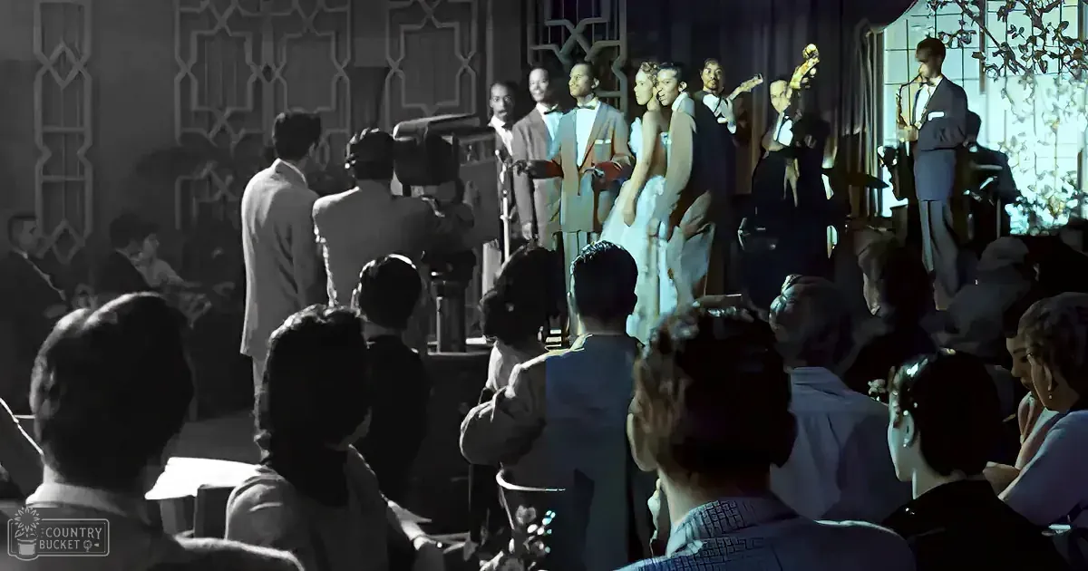 A bride and groom stand on stage, surrounded by musicians and an audience, in a dimly lit, elegant setting.