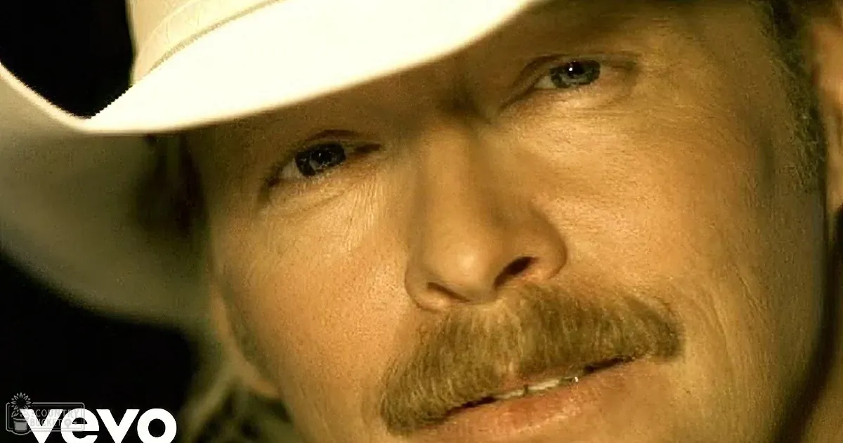 An older adult male in a performance setting, wearing a white cowboy hat and light blue button-up shirt, holding a microphone against a bright green stage backdrop.
