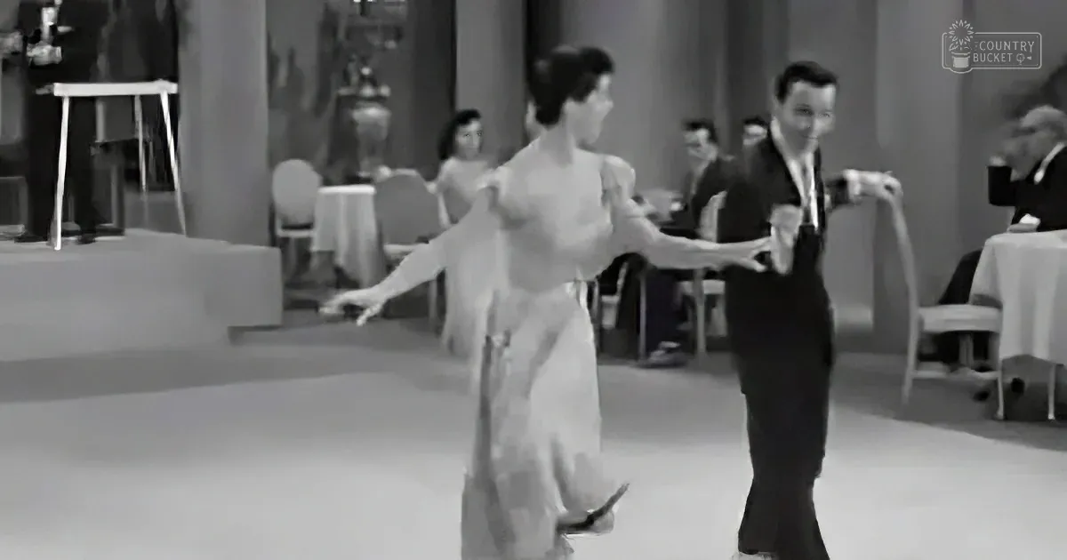 A couple in formal attire dances gracefully on a dance floor, surrounded by elegantly dressed onlookers.
