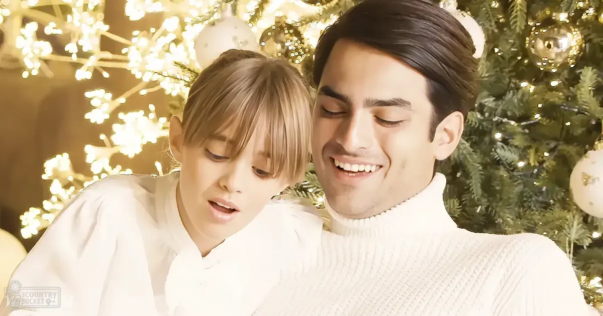 A couple in white sweaters smiles near a Christmas tree adorned with lights and ornaments.