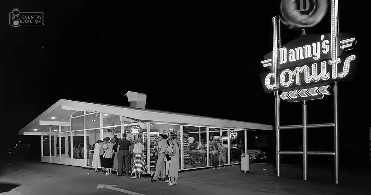 Danny's Donuts neon sign glows brightly at night, attracting a crowd of people outside the illuminated storefront.