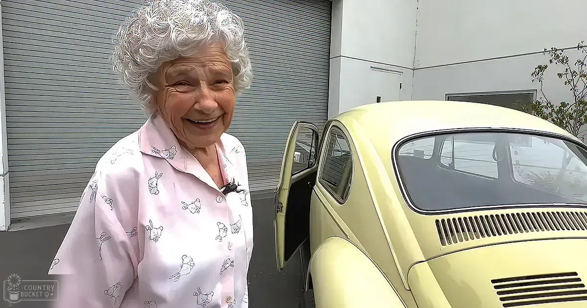 An elderly woman in a pink pajama top smiles near a vintage yellow Volkswagen Beetle with its door open.