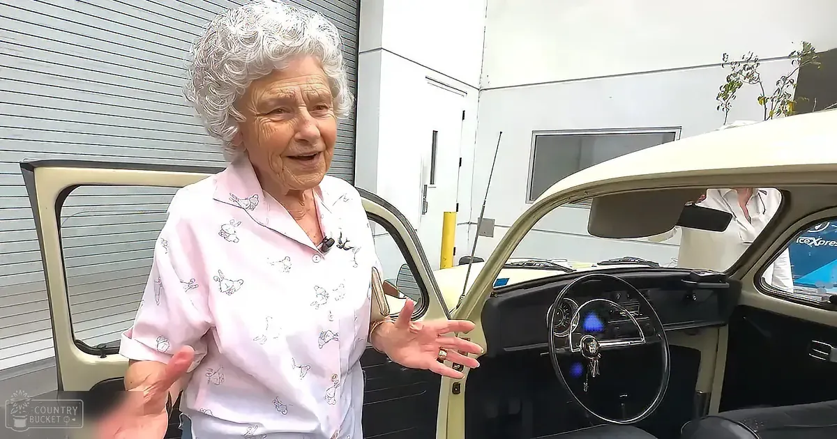 An elderly woman in a pink shirt stands next to a vintage car, gesturing with her hands.