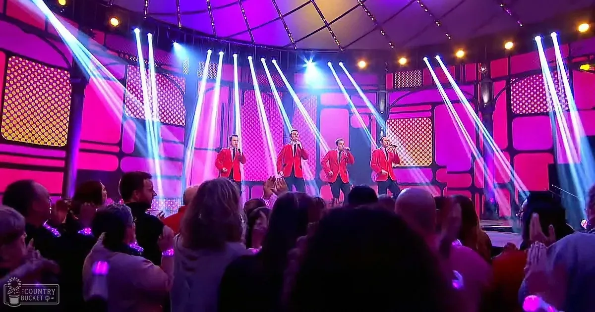 Four men in red suits sing on a stage with bright blue and purple lights, surrounded by an enthusiastic audience.