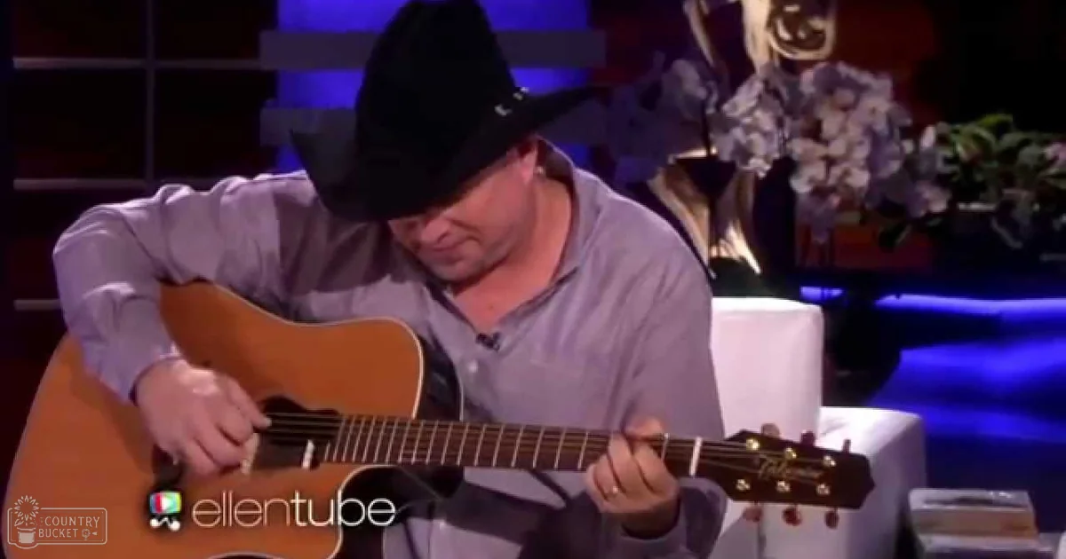 Adult male singer Garth Brooks wearing a black cowboy hat and light gray button-up shirt performing on stage at an Ellen show, with blue stage lighting and floral decor in the background.