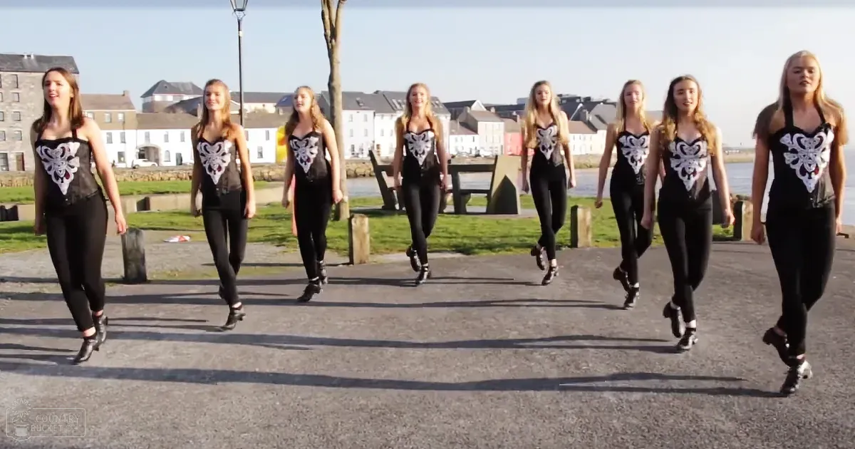 Young adult women performing outdoors, possibly Ireland; spring or summer setting with greenery and a body of water.