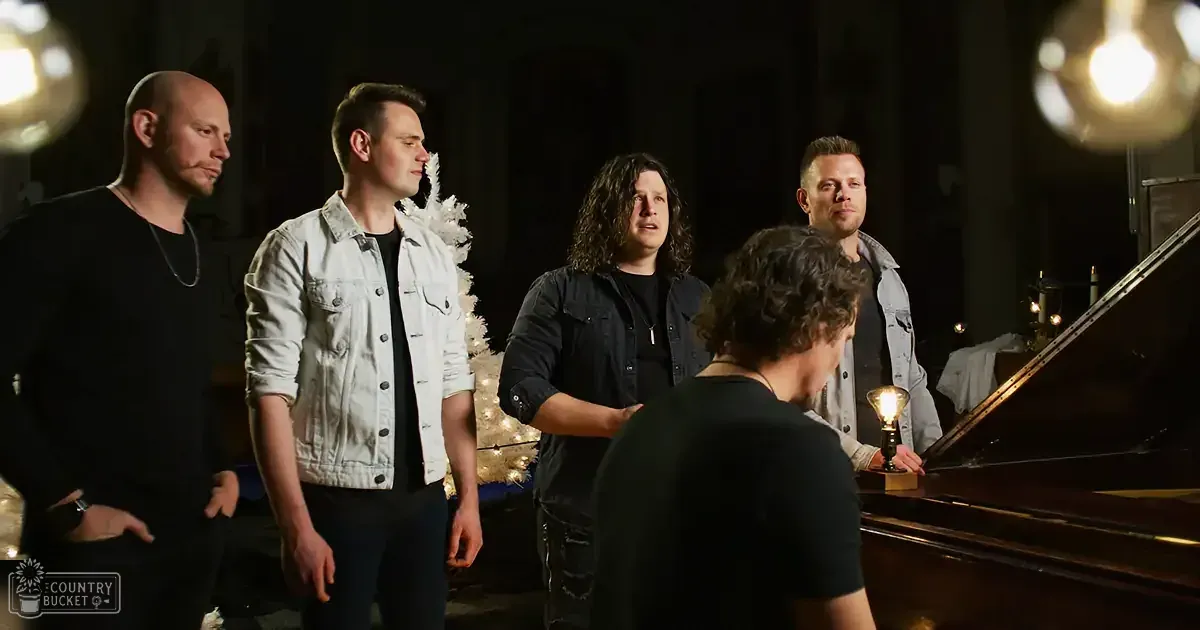 A group of five men stands around a piano in a dimly lit room, with one man playing the piano.