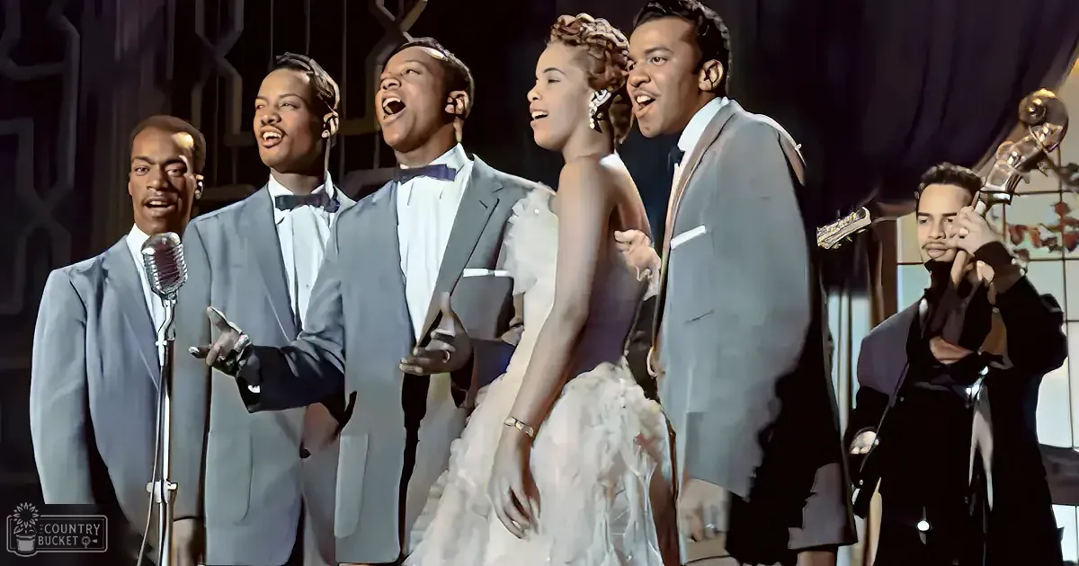A group of five men in suits sing, accompanied by a woman in a white dress and a musician playing a bass.