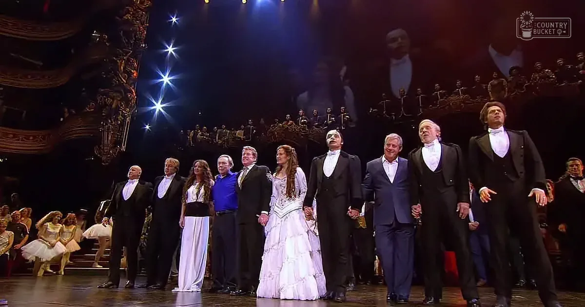 A group of performers in formal attire stands on stage, with dancers in the background, under dramatic lighting.