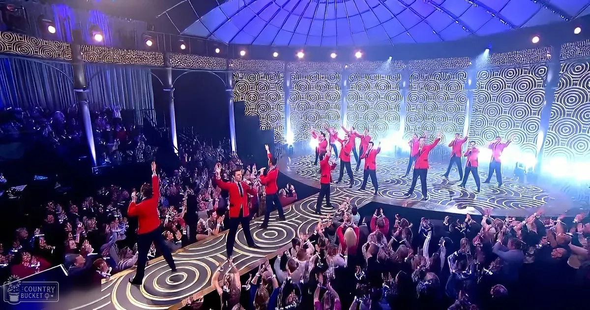 A group in red jackets performs on a stage with a spiral pattern, surrounded by an enthusiastic crowd.