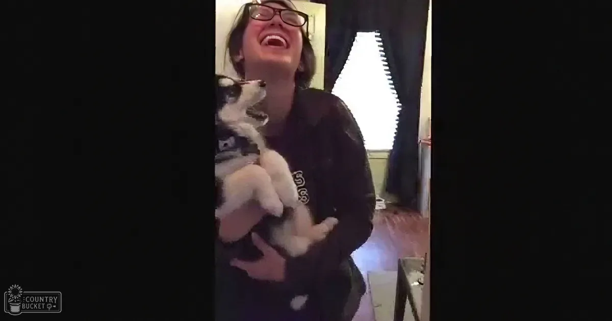 A woman holds a black-and-white puppy, smiling, in a cozy indoor setting.