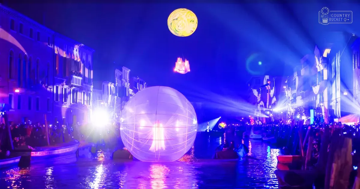 A vibrant nighttime festival scene in a canal city, likely Venice with colorful spotlights and illuminated structures along the waterway.
