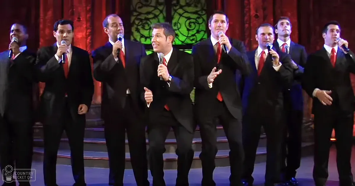 A male a cappella group performing in an indoor setting with red curtains and a green circular design behind them, dressed in dark suits and white shirts with red ties.