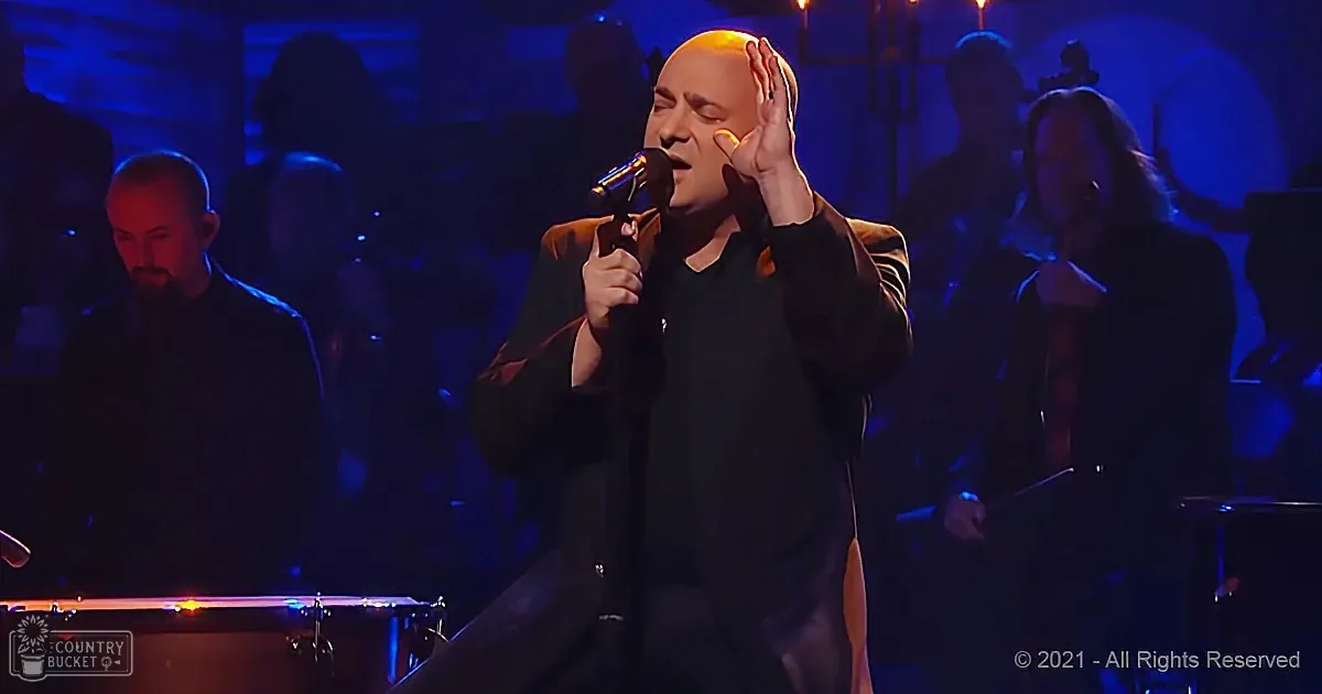 A man in black holds a microphone, singing with his hand on his head, surrounded by musicians in a dimly lit stage setting.