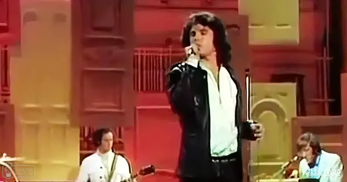 A man in a black leather jacket and white shirt sings into a microphone on stage.