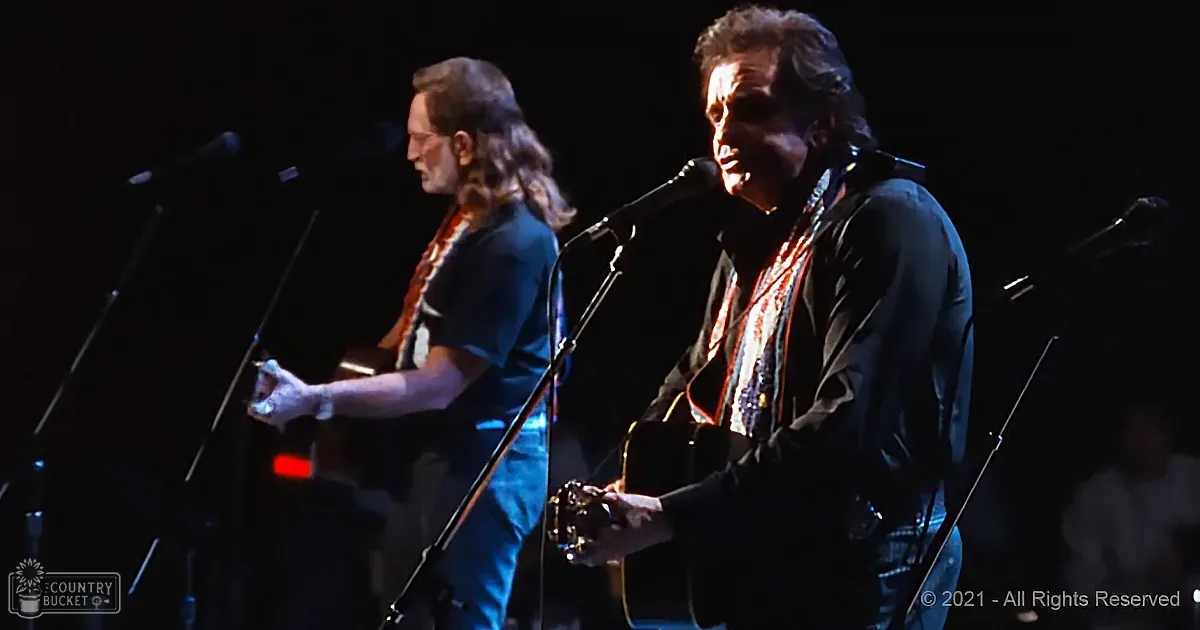 A man in a black shirt and jeans plays guitar on stage with a microphone, another musician in the background.