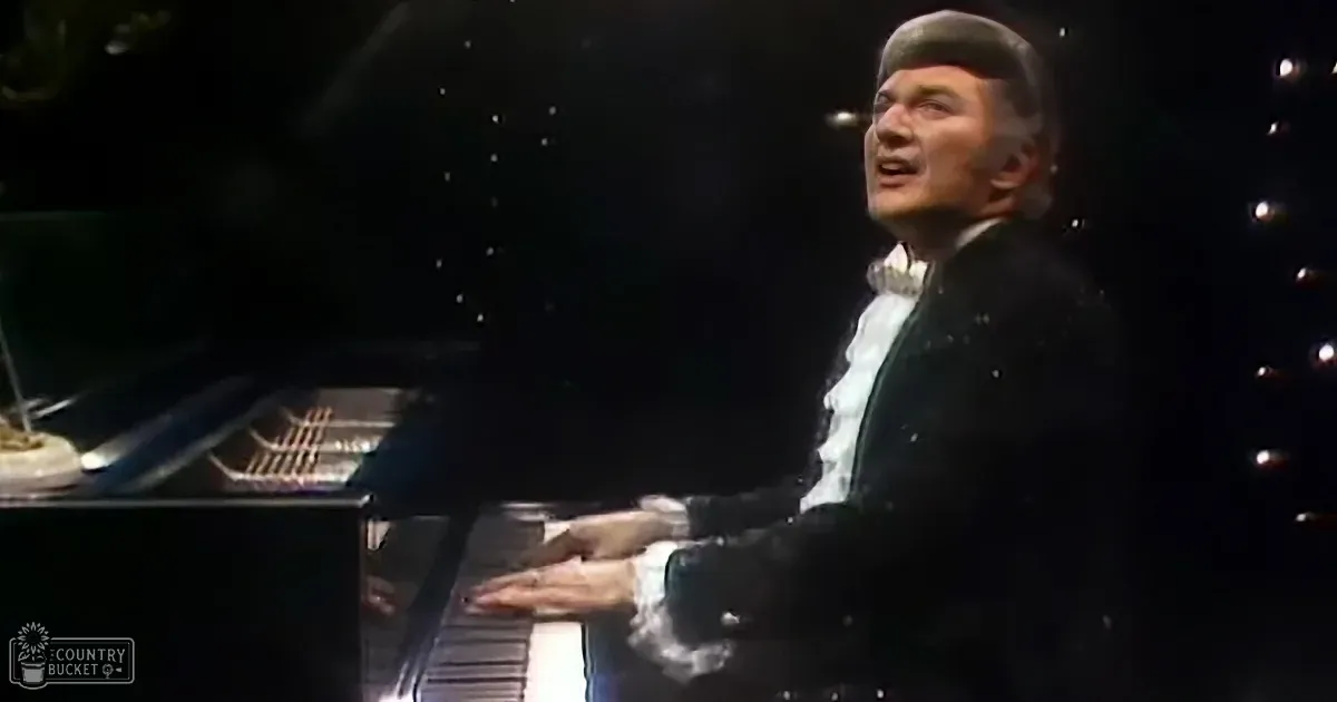 A man in a black suit and hat sings while playing a piano, surrounded by dark background.