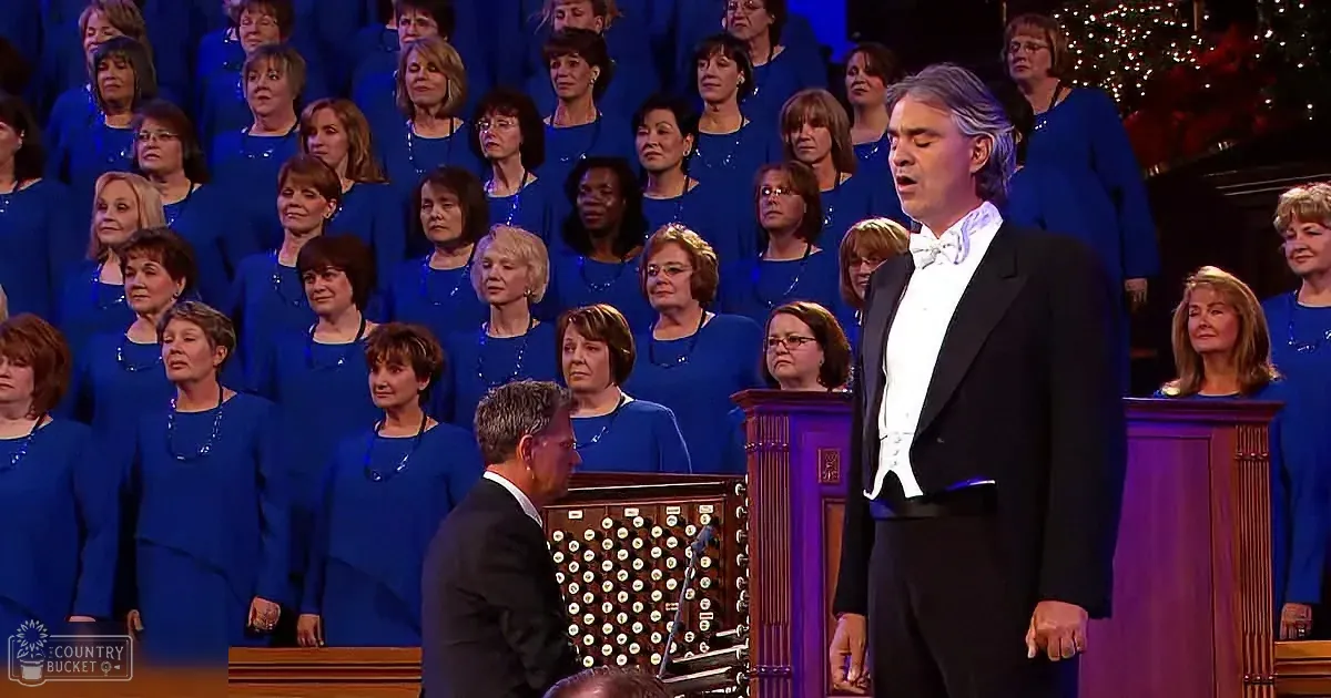 A man in a black suit sings in front of a choir in blue robes, with an organist seated at a pipe organ.