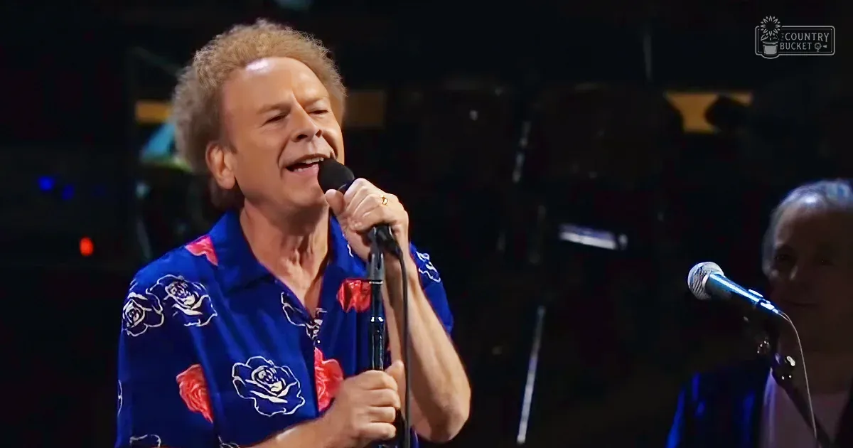A middle-aged man wearing a loose-fitting blue Hawaiian-style shirt with red and white floral patterns performs on stage at an indoor concert venue under bright stage lighting.
