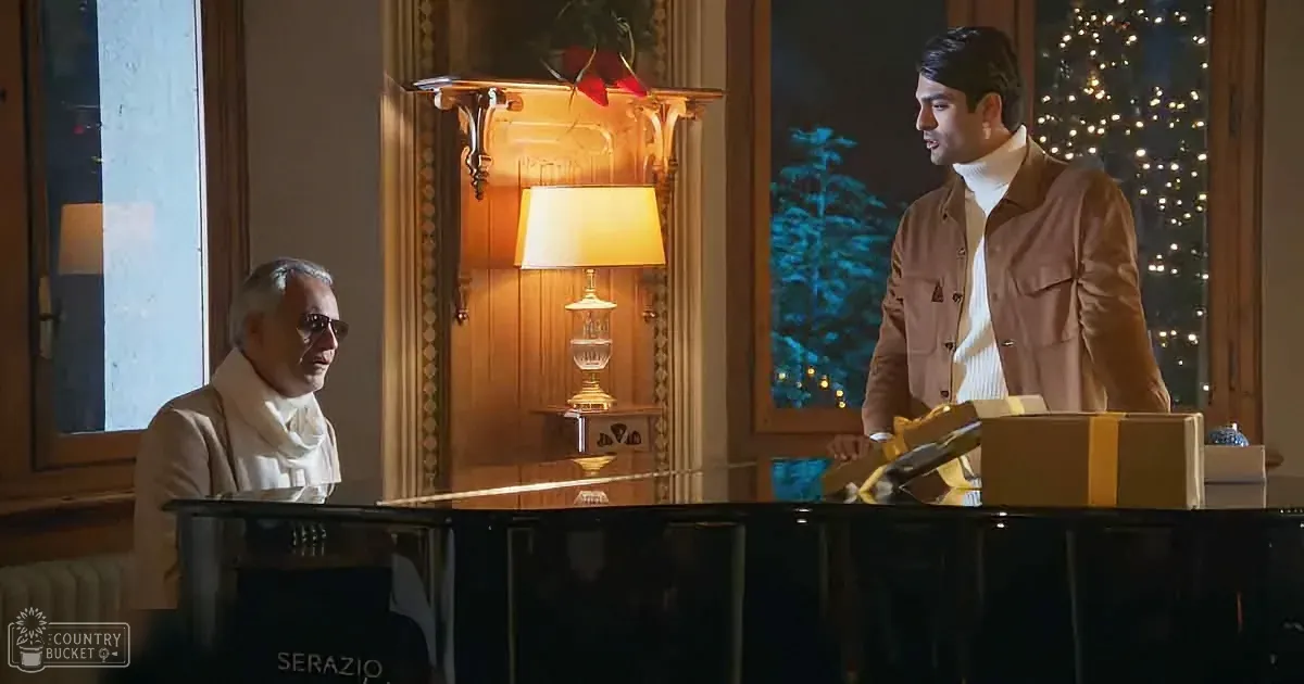 A man in a brown jacket stands beside a piano, holding a gift, while another man in a white scarf sits at the piano.