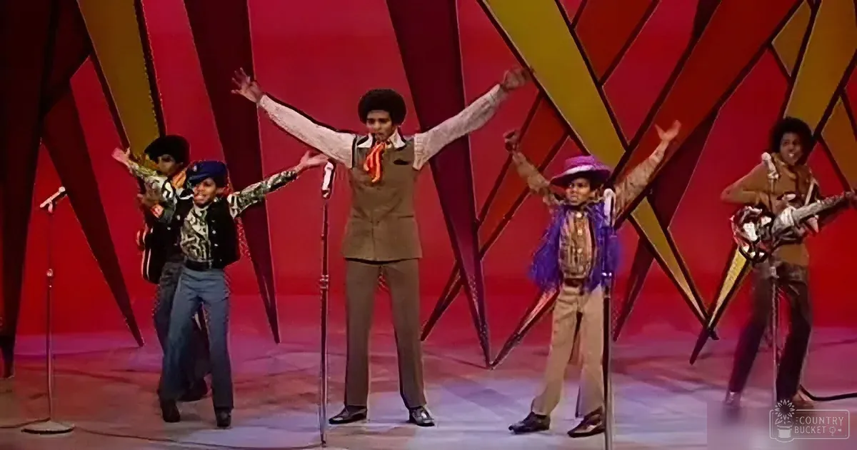 A man in a brown outfit with arms raised, surrounded by other performers in colorful costumes on a stage with a red and...