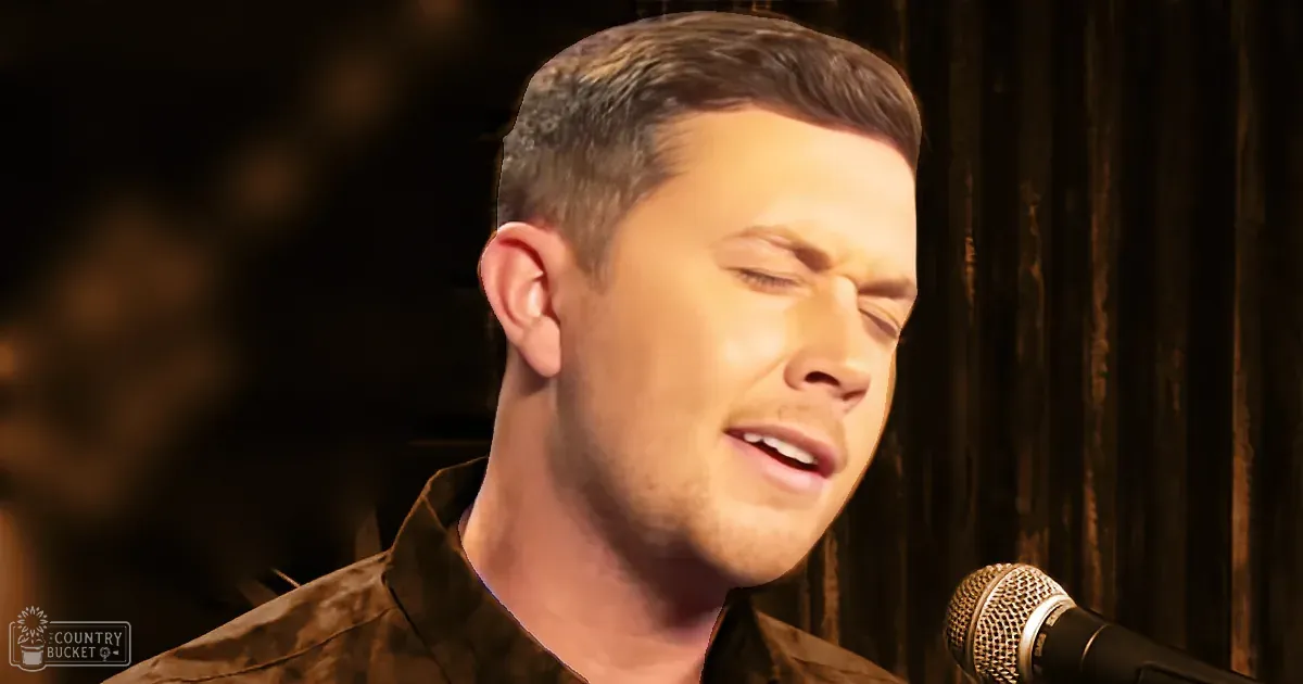 A young adult male singer in a relaxed fit brown collared shirt performs into a microphone against a backdrop of curtains, suggesting an indoor performance or recording session.