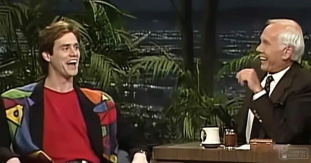 A man in a colorful jacket laughs alongside another man in a suit on a talk show set.