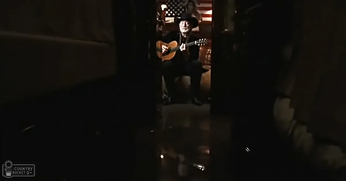 A man in a cowboy hat plays guitar in a dimly lit room with an American flag in the background.