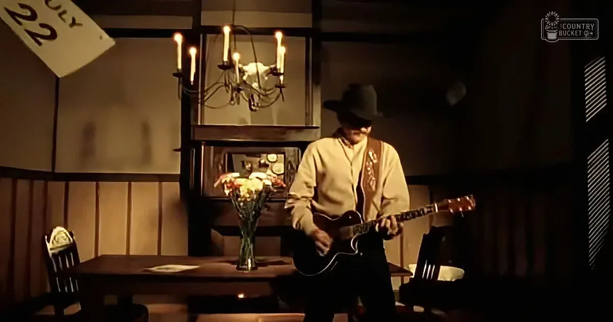 A man in a cowboy hat plays guitar in a dimly lit room with a chandelier and flowers.