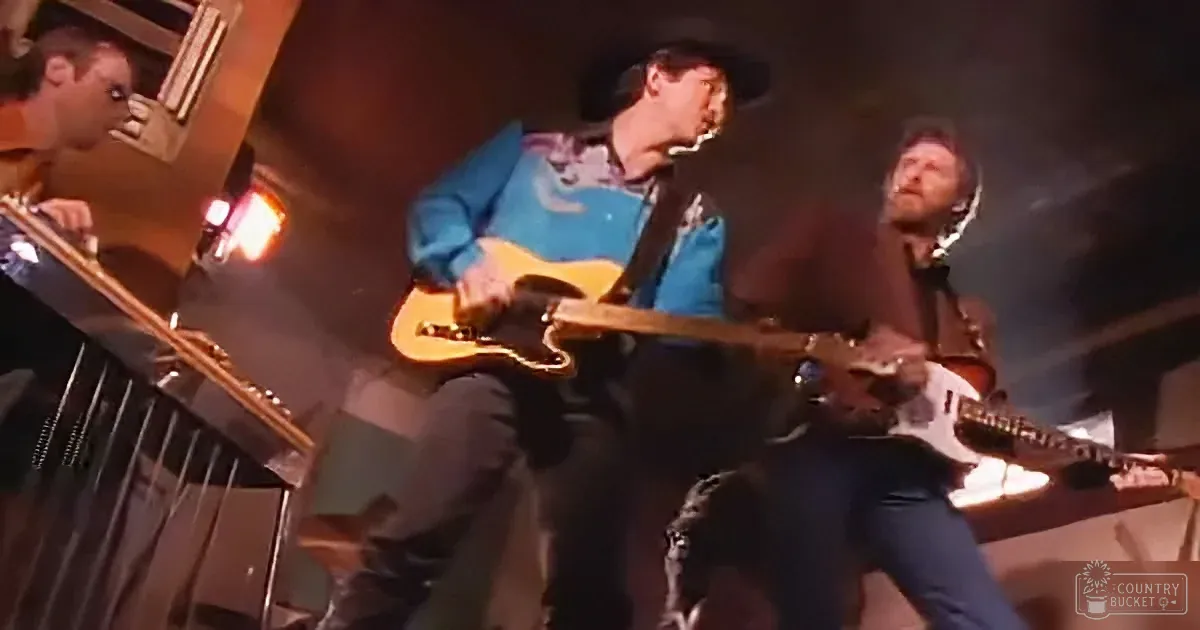A man in a cowboy hat plays a guitar on stage, surrounded by other musicians.