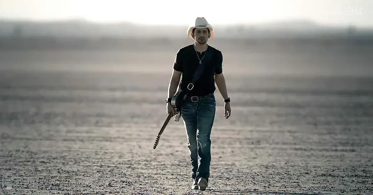 A man in a cowboy hat walks across a vast, barren landscape carrying a guitar.