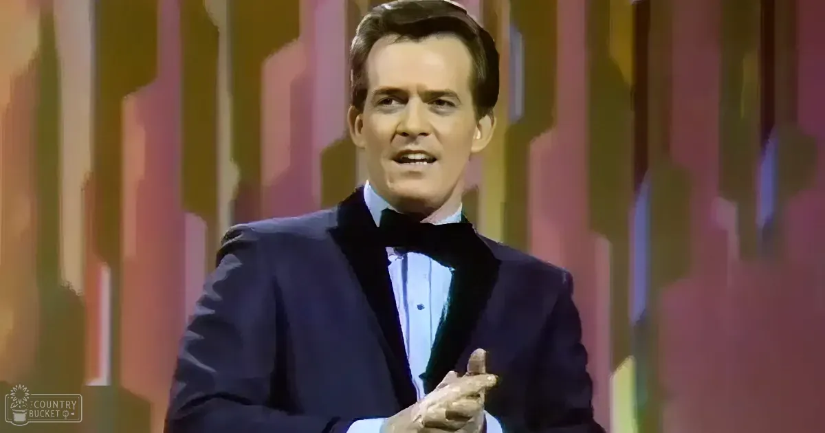 A man in a dark suit and bow tie gestures while speaking on stage.