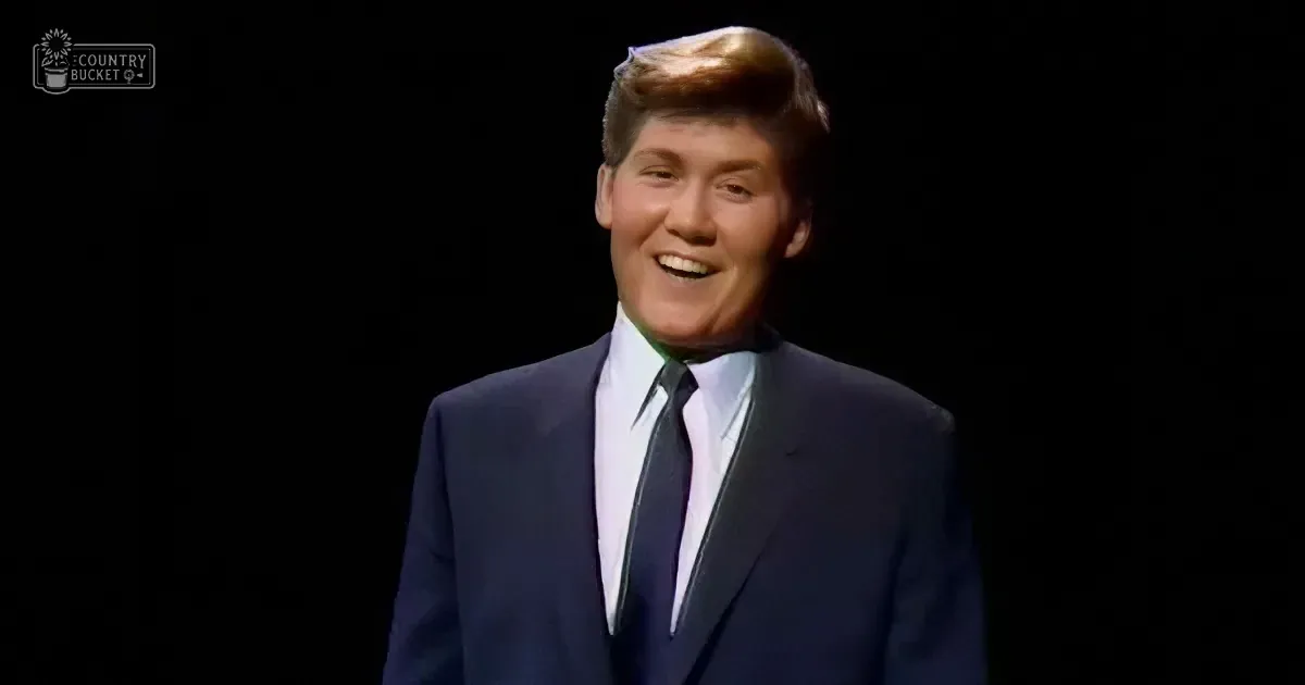 A man in a dark suit and tie smiles brightly against a black background.