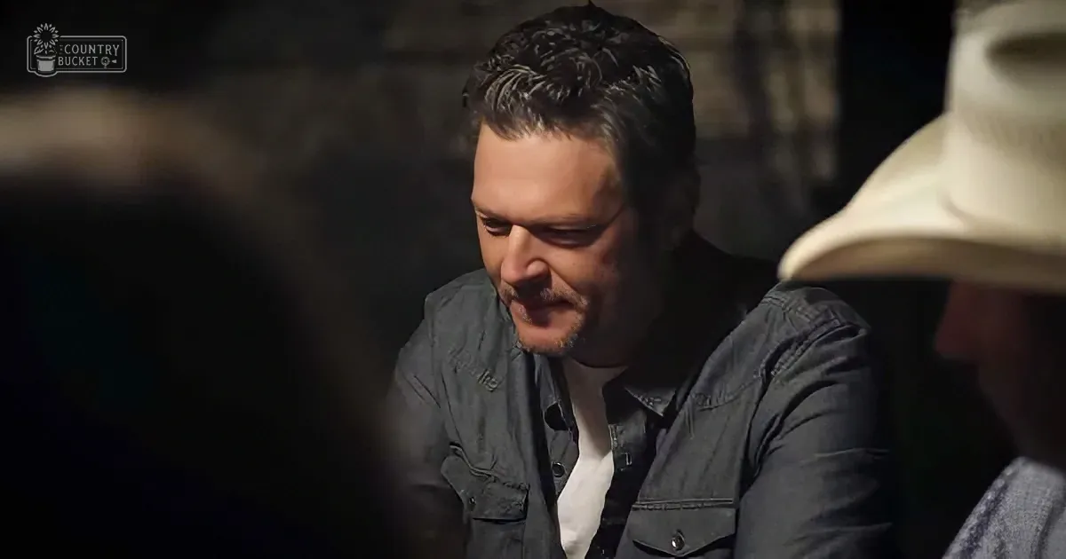 A man in a denim shirt and white shirt is smiling, looking down, with another person in a hat partially visible.