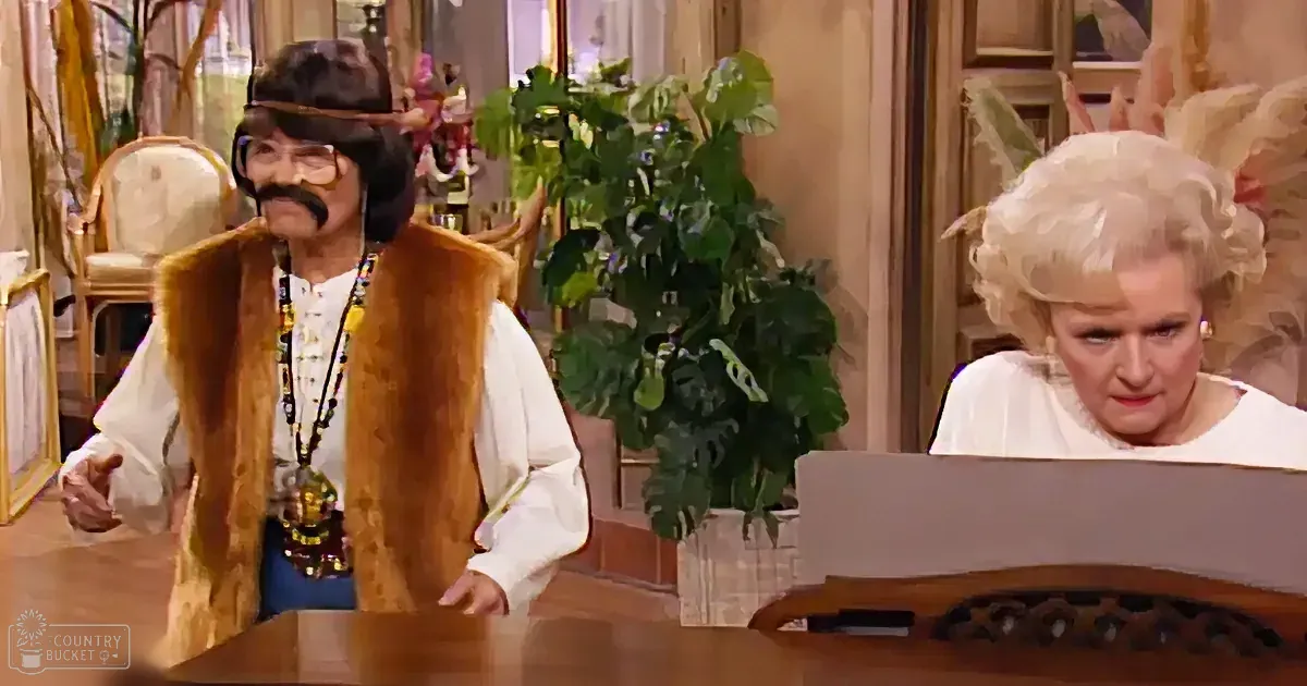 A man in a fur vest and wig stands beside a seated woman in a white outfit, both in a room with plants and furniture.