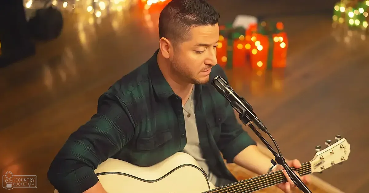 A man in a green plaid shirt plays an acoustic guitar and sings into a microphone.