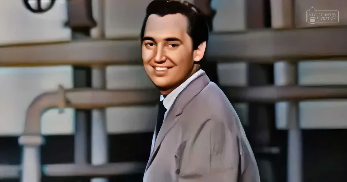 A man in a light gray suit and tie smiles, standing in front of a blurred background with shelves.